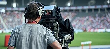Kameramann filmt mit einer Broadcast-Kamera ein Spiel in einem Stadion, von hinten fotografiert.