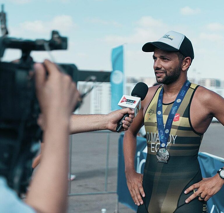 Sportler mit Medaille wird nach einem Wettkampf mit Mikrofon und Kamera interviewt.